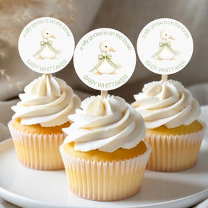 May include: Three cupcakes topped with white frosting and circular paper toppers. Each topper features a watercolor illustration of a white goose wearing a green bow, with the text a silly goose is on the loose and BABY WHITTAKER.