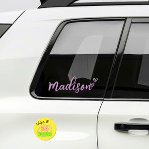May include: A light purple vinyl decal applied to a car window featuring the name Madison in a cursive script font with two small heart outlines positioned to the right of the text. The decal is applied to the glass of a white vehicle.