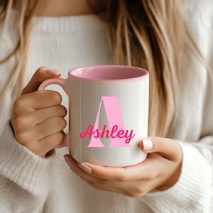 May include: A white ceramic mug with a light pink interior and handle. The front of the mug features a large, light pink capital letter A with the name Ashley printed in a bold, dark pink script font layered over the center of the letter.