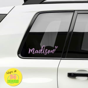 May include: A light purple vinyl decal applied to a car window, featuring the name Madison in a cursive script font followed by two small heart outlines. The decal is positioned on the rear passenger side window of a white vehicle.