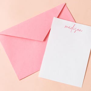 May include: A pink envelope and a white notepad with the name "madison" printed in pink script. The envelope is folded and positioned behind the notepad. The background is a light peach color.