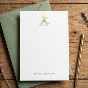 May include: A white rectangular stationery card featuring a watercolour illustration of a small duck wearing a green bow at the top centre and the text BABY MARTINEZ printed in green at the bottom, enclosed by a thin green border line.
