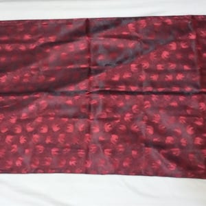 May include: A burgundy scarf with a repeating pattern of small, red floral shapes. The scarf has a fringed edge and is made of a silky material. The scarf is rectangular and appears to be folded.