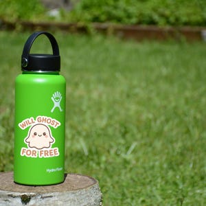 May include: A green Hydro Flask water bottle with a black handle and lid. A sticker on the bottle says "Will Ghost For Free" with a cartoon ghost illustration. The bottle is sitting on a wooden stump against a green grass background.