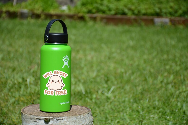 May include: A green Hydro Flask water bottle with a black handle and lid. A sticker on the bottle says "Will Ghost For Free" with a cartoon ghost illustration. The bottle is sitting on a wooden stump against a green grass background.