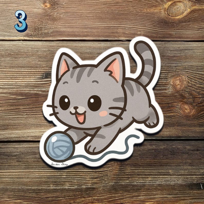 May include: A gray cartoon cat sticker with a playful design. The cat is depicted with a happy expression, playing with a blue ball of yarn. The number "3" is in the upper left corner. The sticker is on a wooden surface.