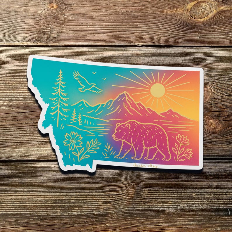 May include: A sticker in the shape of Montana, featuring a colorful landscape illustration. The design includes a bear, mountains, a sun, and an eagle. The color palette transitions from teal to orange and yellow.