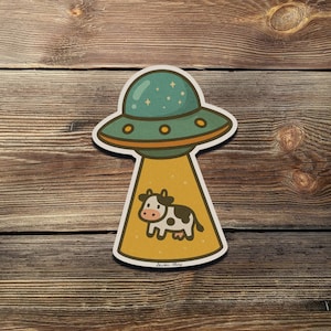 May include: A sticker featuring a cartoon UFO with a teal top and orange accents. A yellow beam shines down on a cow with black and white spots. The sticker is on a wooden background.