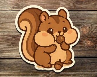 Puede incluir: Una pegatina con una ardilla de dibujos animados en tonos marrones, sosteniendo una nuez en la boca. La ardilla tiene una cola tupida y mejillas rosadas. La pegatina está sobre una superficie de madera.