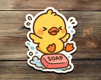 May include: A yellow cartoon duck sticker with orange feet and a beak, splashing in blue water. The duck is standing on a pink bar of soap with the word "SOAP" printed on it. The sticker is on a wooden background.