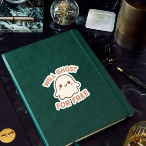 May include: A dark green journal with a matching ribbon bookmark. A sticker on the cover reads "Will Ghost For Free" with a cartoon ghost illustration. The journal is on a black marble surface, surrounded by office supplies.