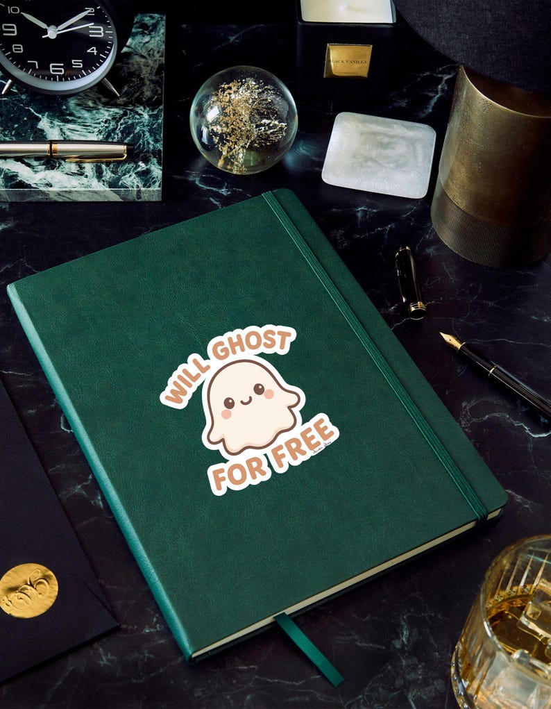 May include: A dark green journal with a matching ribbon bookmark. A sticker on the cover reads "Will Ghost For Free" with a cartoon ghost illustration. The journal is on a black marble surface, surrounded by office supplies.