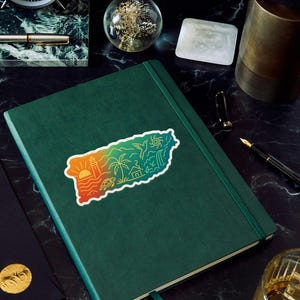 May include: A green leather-bound journal with a colorful sticker depicting a coastal landscape, including a lighthouse, palm trees, and a turtle. The journal is on a dark, marbled surface, with a pen, clock, and other decorative items.
