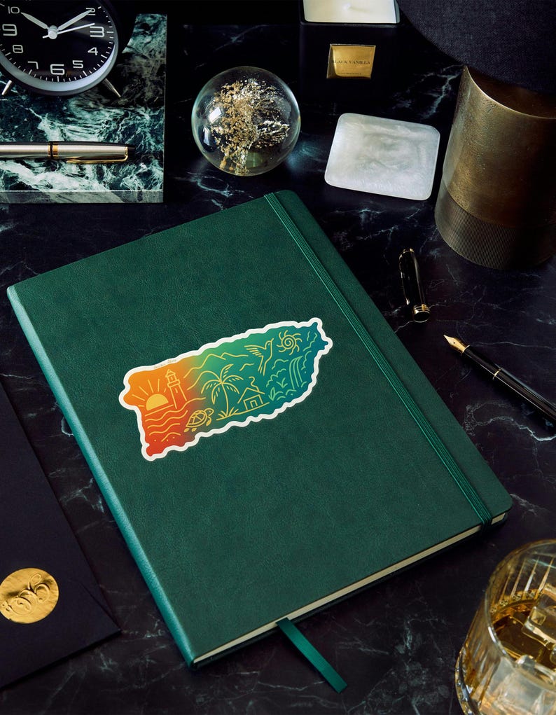 May include: A green leather-bound journal with a colorful sticker depicting a coastal landscape, including a lighthouse, palm trees, and a turtle. The journal is on a dark, marbled surface, with a pen, clock, and other decorative items.