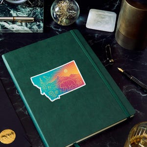 May include: A green journal with a colorful sticker of the state of Montana, featuring a bear, mountains, and a sun. The journal is on a dark, patterned surface, with a pen, clock, and other desk accessories.
