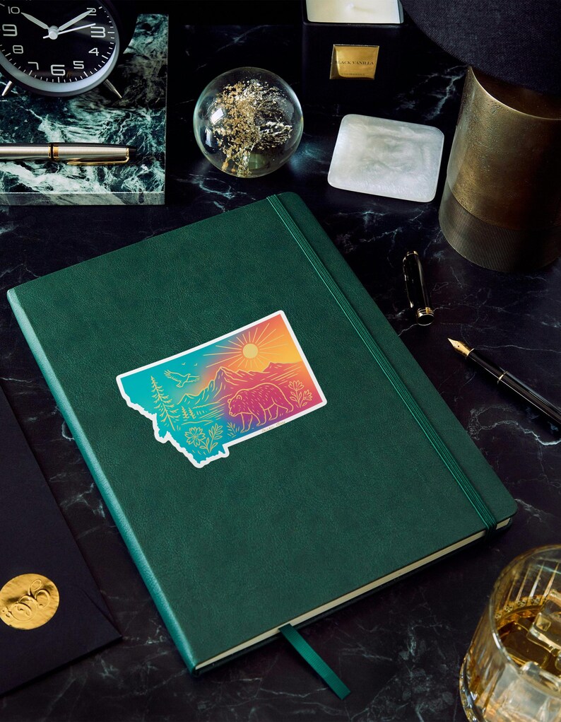 May include: A green journal with a colorful sticker of the state of Montana, featuring a bear, mountains, and a sun. The journal is on a dark, patterned surface, with a pen, clock, and other desk accessories.