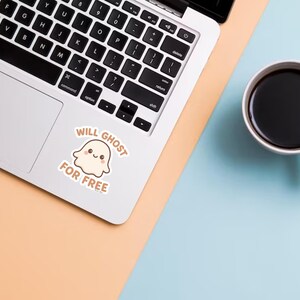 May include: A laptop with a sticker that says "Will Ghost For Free" next to a cup of coffee. The sticker features a cute ghost illustration. The laptop is on a peach and blue surface.