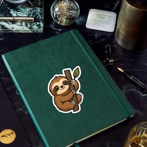 May include: A green journal with a sloth sticker holding onto a branch with a leaf. The sticker is brown and white. The journal has a green ribbon bookmark. Other items on the surface include a clock, pen, and a glass sphere.