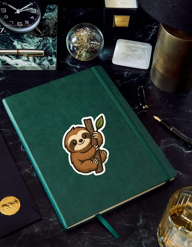 May include: A green journal with a sloth sticker holding onto a branch with a leaf. The sticker is brown and white. The journal has a green ribbon bookmark. Other items on the surface include a clock, pen, and a glass sphere.