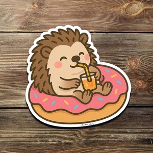 Puede incluir: Un erizo de dibujos animados con un donut rosa y blanco con chispas, sosteniendo un vaso de zumo de naranja con una pajita. El erizo sonríe y tiene una expresión feliz.