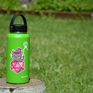 Może przedstawiać: Zielona butelka na wodę Hydro Flask z czarnym uchwytem i pokrywką. Naklejka na butelce przedstawia kresk&oacute;wkowego kota w kapeluszu czarodzieja, siedzącego na r&oacute;żowo-białej kostce D20. Na kostce widnieje liczba 20.
