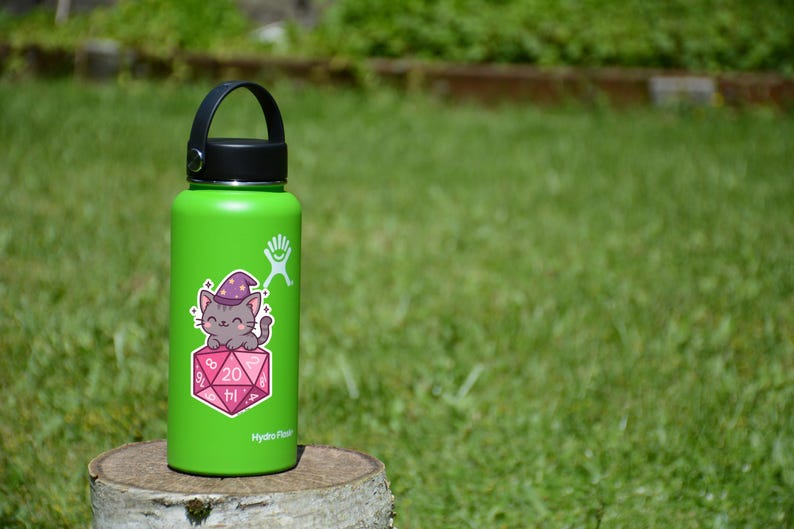 Może przedstawiać: Zielona butelka na wodę Hydro Flask z czarnym uchwytem i pokrywką. Naklejka na butelce przedstawia kresk&oacute;wkowego kota w kapeluszu czarodzieja, siedzącego na r&oacute;żowo-białej kostce D20. Na kostce widnieje liczba 20.