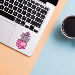 Może przedstawiać: Laptop z naklejką przedstawiającą szarego kota z kresk&oacute;wki w fioletowym kapeluszu czarodzieja, siedzącego na r&oacute;żowej dwudziestościennej kostce. Biały kubek wypełniony czarną kawą stoi na niebieskiej powierzchni.