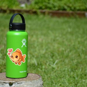 May include: A green insulated water bottle with a black handle and lid. A sticker of a cartoon goldfish with hearts is on the bottle. The bottle is sitting on a tree stump in front of a green background.