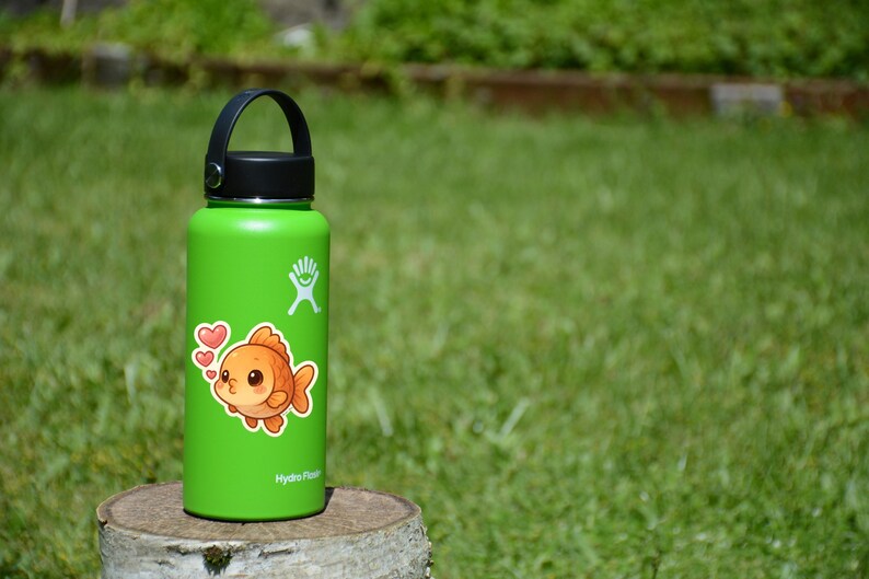 May include: A green insulated water bottle with a black handle and lid. A sticker of a cartoon goldfish with hearts is on the bottle. The bottle is sitting on a tree stump in front of a green background.