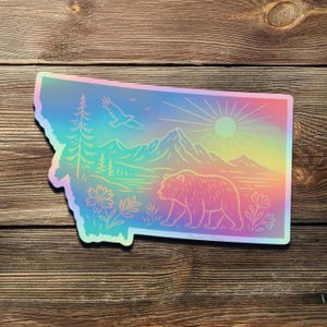 May include: A holographic sticker in the shape of Montana, featuring a bear, mountains, trees, and an eagle. The design has a rainbow gradient and a sunburst.