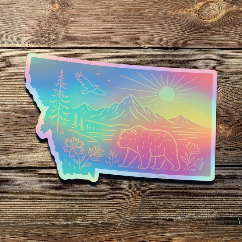 May include: A holographic sticker in the shape of Montana, featuring a bear, mountains, trees, and an eagle. The design has a rainbow gradient and a sunburst.