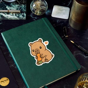May include: A green journal with a sticker of a brown cartoon wombat eating a carrot. The sticker has a small orange heart. The journal is on a black marble surface with a clock, pen, and other items.