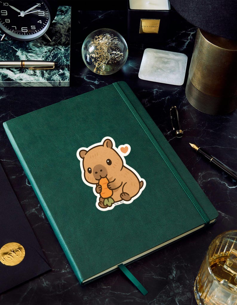 May include: A green journal with a sticker of a brown cartoon wombat eating a carrot. The sticker has a small orange heart. The journal is on a black marble surface with a clock, pen, and other items.