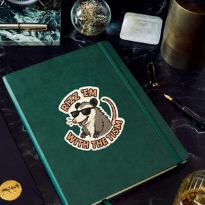 May include: A dark green journal with a sticker that says "Rizz 'em with the tism" featuring a cartoon opossum wearing sunglasses. The journal has a matching green ribbon bookmark and is on a black marble surface.