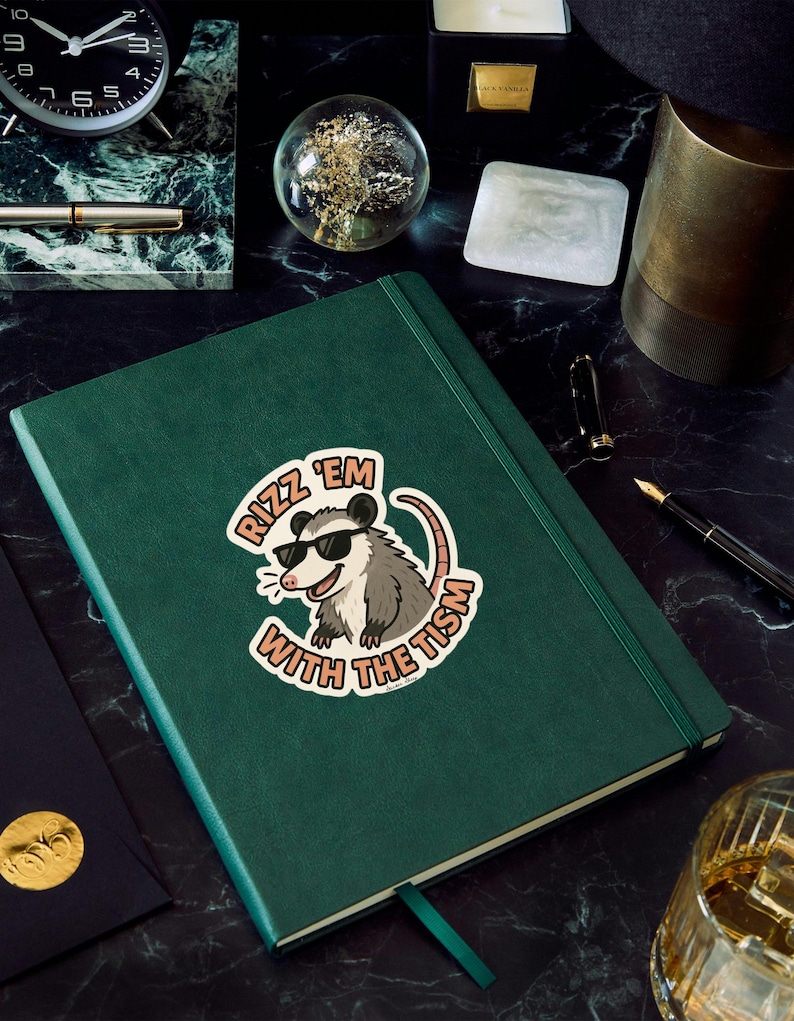 May include: A dark green journal with a sticker that says "Rizz 'em with the tism" featuring a cartoon opossum wearing sunglasses. The journal has a matching green ribbon bookmark and is on a black marble surface.