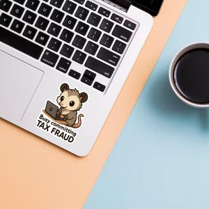 K&ouml;nnte beinhalten: Ein Laptop mit einem Aufkleber eines Cartoon-Opossums, das an einem Laptop arbeitet, mit dem Text "Busy committing TAX FRAUD". Eine wei&szlig;e Tasse mit schwarzem Kaffee steht auf einer hellblauen Oberfl&auml;che.