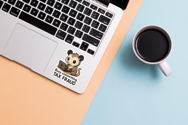 K&ouml;nnte beinhalten: Ein Laptop mit einem Aufkleber eines Cartoon-Opossums, das an einem Laptop arbeitet, mit dem Text "Busy committing TAX FRAUD". Eine wei&szlig;e Tasse mit schwarzem Kaffee steht auf einer hellblauen Oberfl&auml;che.