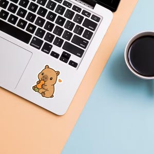 May include: A sticker of a cartoon wombat eating a carrot is on a laptop. The wombat is brown with a small heart next to it. A white mug filled with black coffee sits on a blue and peach surface.