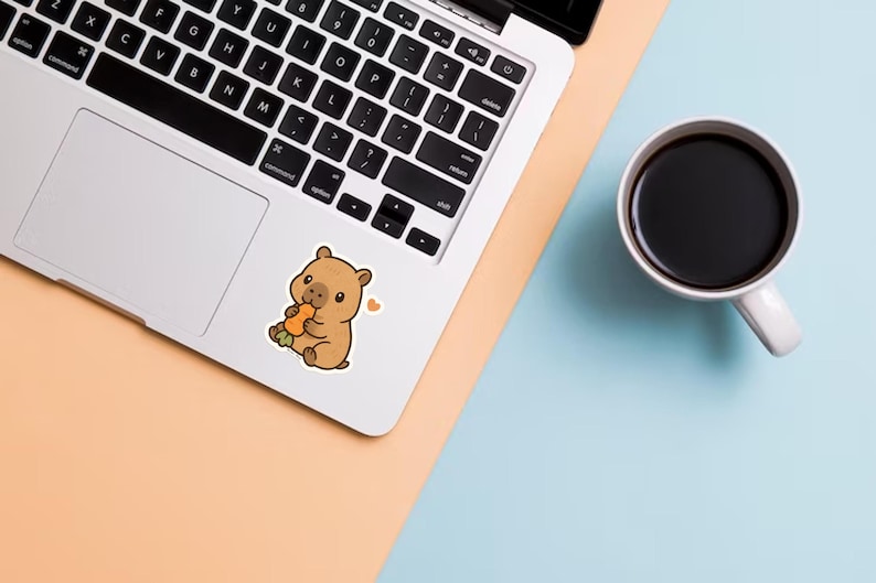 May include: A sticker of a cartoon wombat eating a carrot is on a laptop. The wombat is brown with a small heart next to it. A white mug filled with black coffee sits on a blue and peach surface.