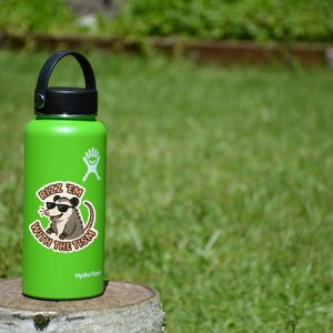 May include: A green Hydro Flask water bottle with a black handle and lid. A sticker on the bottle features a cartoon opossum wearing sunglasses and the text "Rizz 'em with the Tism."