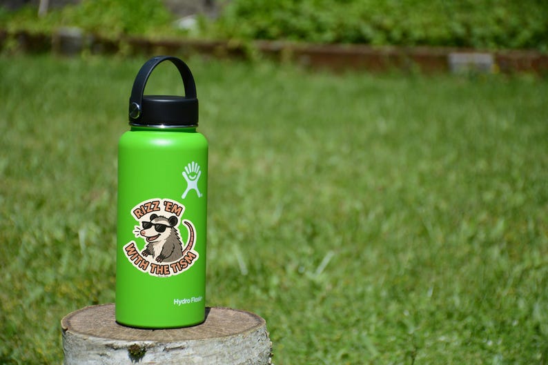 May include: A green Hydro Flask water bottle with a black handle and lid. A sticker on the bottle features a cartoon opossum wearing sunglasses and the text "Rizz 'em with the Tism."