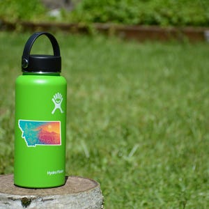 May include: A green Hydro Flask water bottle with a black lid and handle. A sticker of the state of Montana with a colorful landscape is affixed to the bottle. The bottle is sitting on a tree stump in a grassy area.