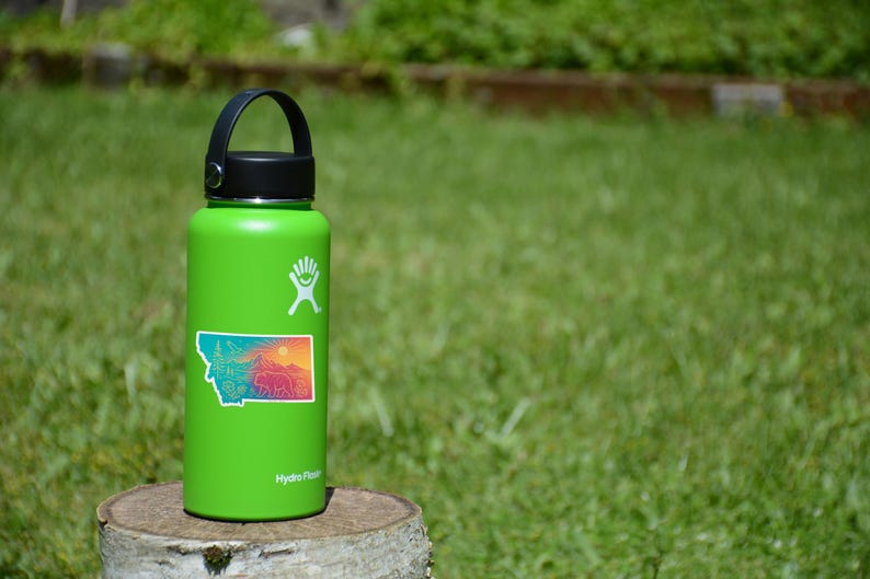 May include: A green Hydro Flask water bottle with a black lid and handle. A sticker of the state of Montana with a colorful landscape is affixed to the bottle. The bottle is sitting on a tree stump in a grassy area.