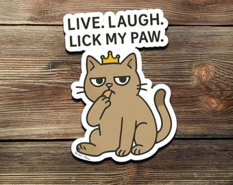 Può includere: Un adesivo con un gatto dei cartoni animati con una corona, che si lecca la zampa. Il testo sopra recita "LIVE. LAUGH. LICK MY PAW." Il gatto è marrone con un contorno bianco, su sfondo di legno.
