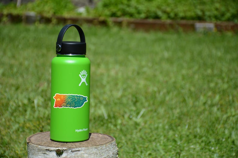 May include: A green Hydro Flask water bottle with a black lid and handle. A colorful sticker of a map of Puerto Rico is on the front. The bottle is sitting on a tree stump in front of a green grassy background.