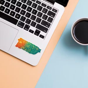 May include: A laptop with a colorful sticker of Puerto Rico, featuring a sunset, palm trees, and ocean waves. A white mug filled with black coffee sits on a light blue surface.