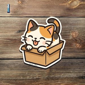 May include: A die-cut sticker featuring a cartoon calico cat with a happy expression, sticking its tongue out while sitting in a cardboard box. The sticker has a white border and is set against a wooden background.