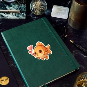May include: A green journal with a sticker of a cute orange goldfish with pink hearts. The journal is on a black marble surface, with a clock, pen, and other desk accessories.