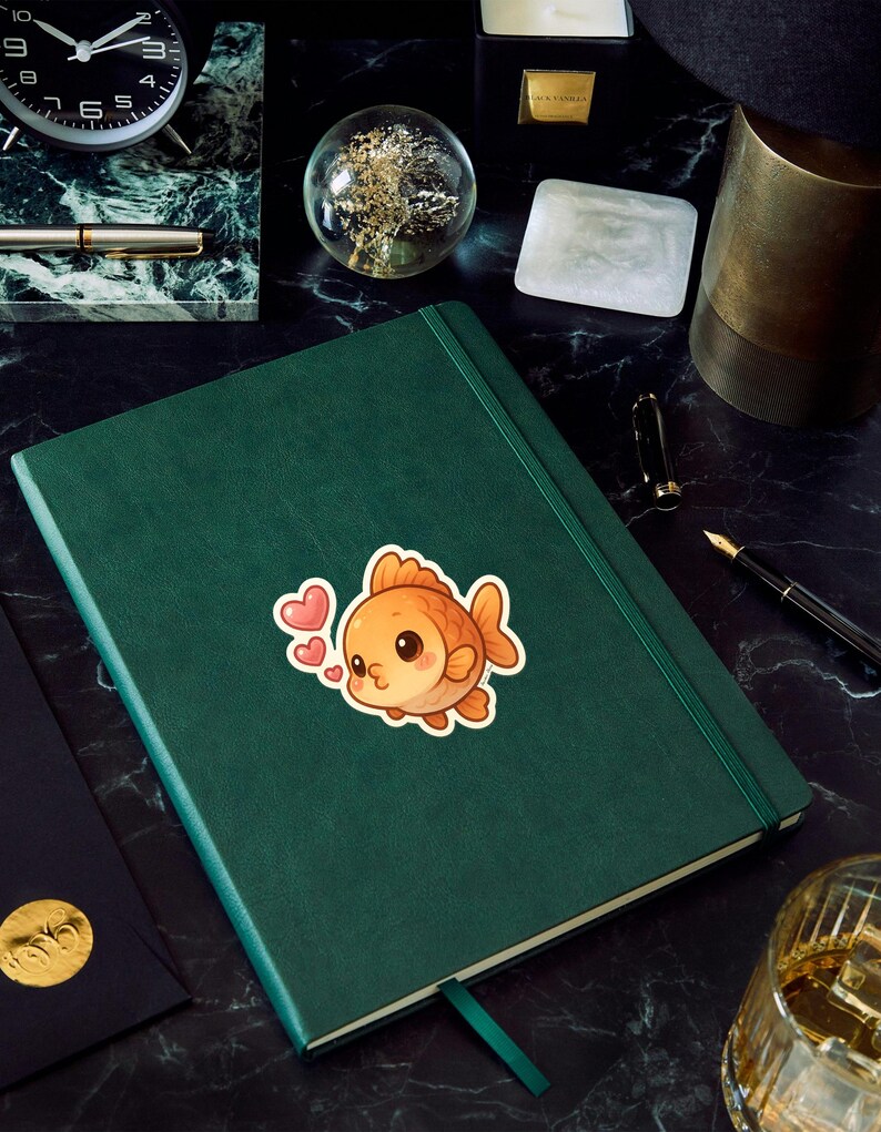 May include: A green journal with a sticker of a cute orange goldfish with pink hearts. The journal is on a black marble surface, with a clock, pen, and other desk accessories.