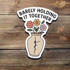 Op de afbeelding: Een sticker met een gebarsten vaas en bloemen, met de tekst "Barely Holding It Together".